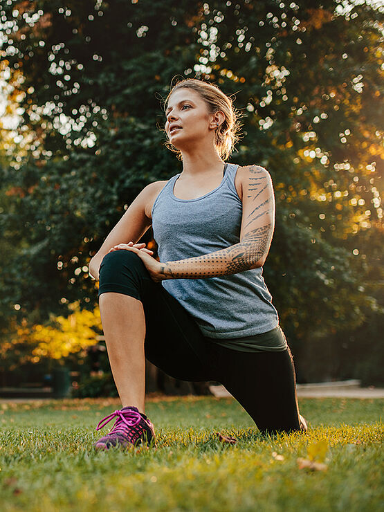 A woman in athletic wear stretches in a park with the sun shining through the trees.